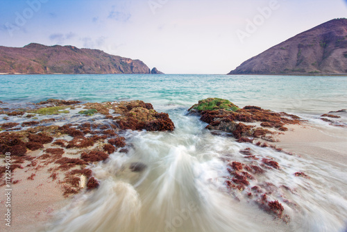 view of the vast blue sea with white waves hitting the coastline containing brown seaweed with two large hills in the middle of the sea and blue skies on Mawun Beach photo