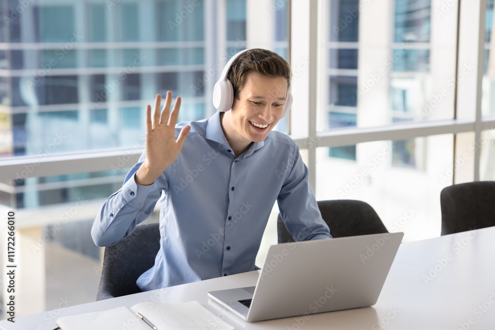 Happy young project manager man in wireless headphones speaking on video conference call at laptop, waving hand hello at screen, using Internet, digital gadget for successful business communication