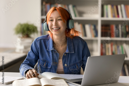 Happy remote student. Smiling trendy looking teen girl spend learning time at college library listen to distant course in earphones write up lecture theses look aside distracted from notebook screen photo