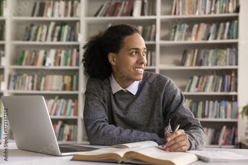 Creative studying. Smiling stylish teenage male learn new material on self training at library look aside distracted from notebook books having idea insight for written work create essay research plan photo