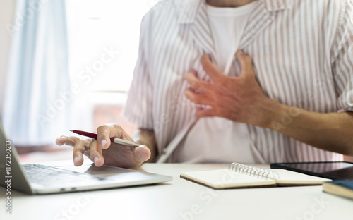 Asian man sitting at desk feel chest pain while using laptop working at home, heart disease exacerbation concept photo