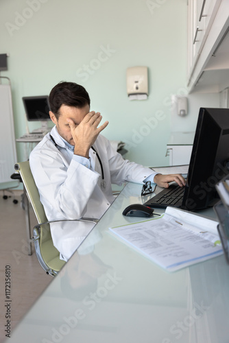 Tired depressed doctor man sitting at workplace with computer in examination room, typing, touching head, suffering from headache, overwork, feeling exhausted, sick, unhappy