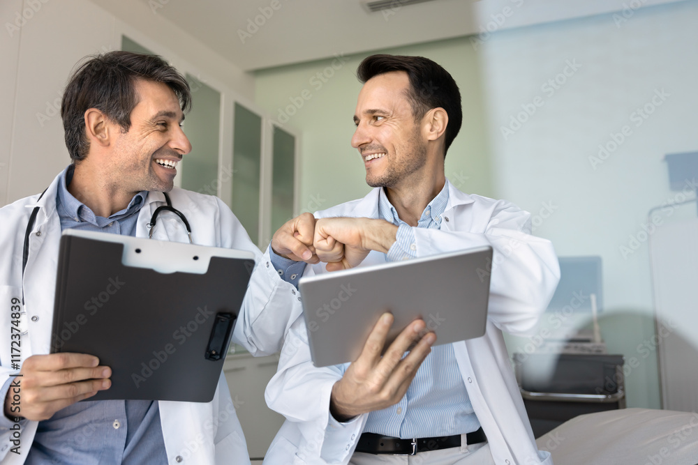 Two happy medical professional friends bumping fists due to successful teamwork, collaboration, holding tablet and paper documents with records, reports, smiling, laughing Two happy medical professional friends bumping fists due to successful teamwork, collaboration, holding tablet and paper documents with records, reports, smiling, laughing
