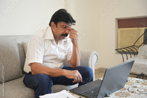 Stressed Mature indian man sitting at home with laptop having pain in eyes after long working hours on computer screen, worried male feeling sad after loss in business.  photo