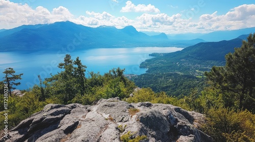 Stunning panoramic view from mountain summit overlooking tranquil lake surrounded by lush greenery and distant hills under blue sky.