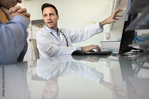 Serious doctor man giving consultation to patient at desktop, pointing at monitor, speaking, explaining examination result, electronic medical reports after checkup procedures photo