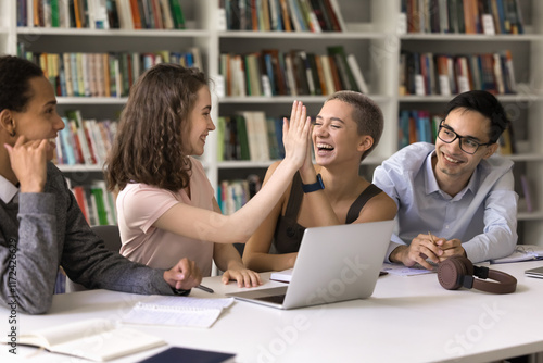 Motivated teamwork. Laughing teen female polyethnic student community member give high five to happy hipster girl acknowledge friend success in research work excited with sudden insight on brainstorm photo