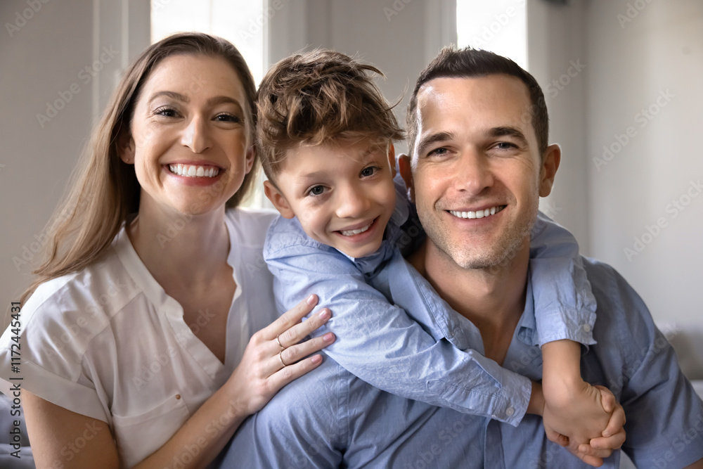 Happy attractive couple of parents and cute son child sitting close together indoors, looking at camera with toothy smiles, enjoying family happiness, togetherness, posing for portrait Happy attractive couple of parents and cute son child sitting close together indoors, looking at camera with toothy smiles, enjoying family happiness, togetherness, posing for portrait