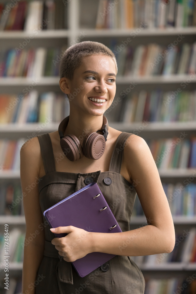 © fizkes - Modern day scientist. Vertical photo portrait happy young lady getting education study work in library stand by bookshelves with folder for papers digital headset device around neck look away dreaming