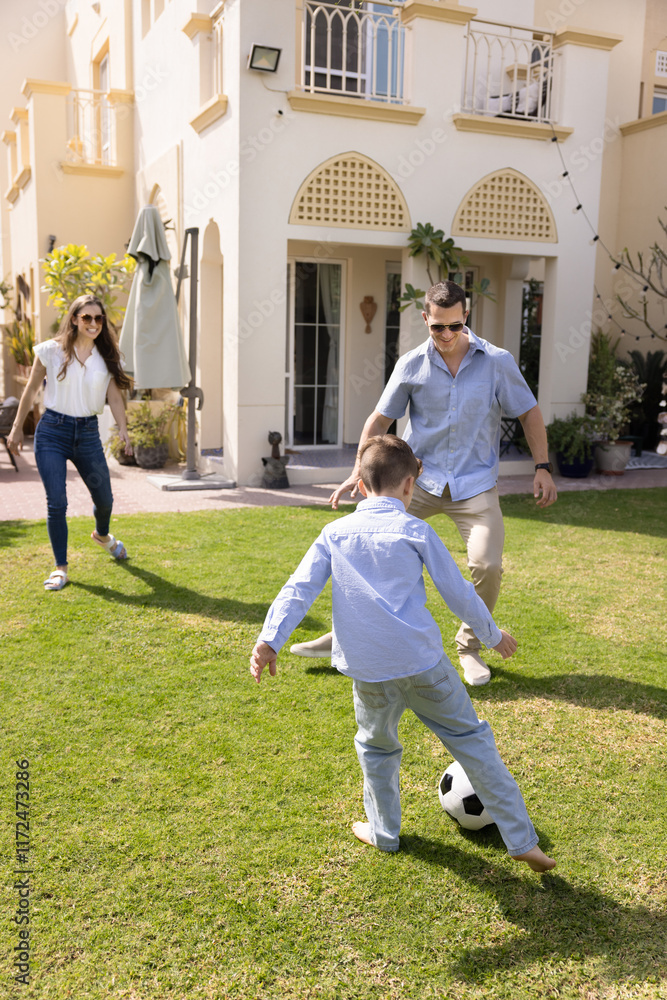 Fototapeta premium Active couple of parents and kid boy playing football on house backyard, kicking ball on lawn, enjoying game, family activity, having fun. Mom and dad training soccer player son outside