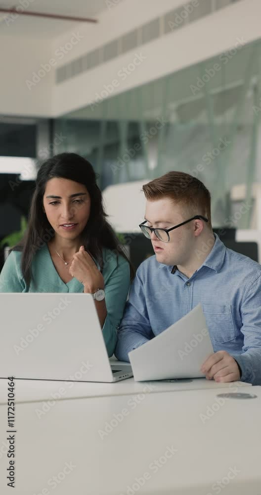 Female mentor provide professional knowledge, share explanation and information about tasks to-do, corporate assignment or software to young 25s man with Down syndrome sit together at desk with laptop Female mentor provide professional knowledge, share explanation and information about tasks to-do, corporate assignment or software to young 25s man with Down syndrome sit together at desk with laptop