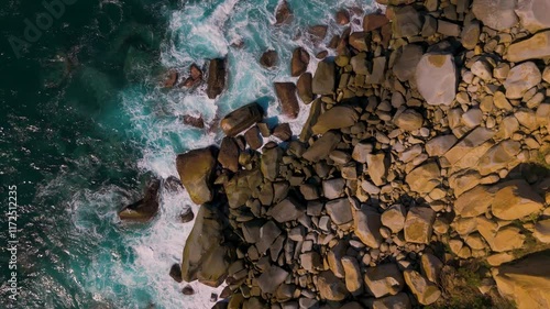 Aerial view of a rocky coastline gently pounded by the waves of the Pacific Ocean. The turquoise blue of the water contrasts with the warm colors of the rocks.