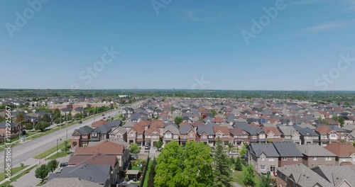 Slow aerial ascent over a suburban neighborhood with rows of detached homes, a main road, and expansive greenery in the distance under clear blue skies.