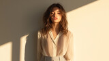 woman with long hair stands against light wall, wearing sheer blouse and high waisted pants, illuminated by soft sunlight, creating serene atmosphere.