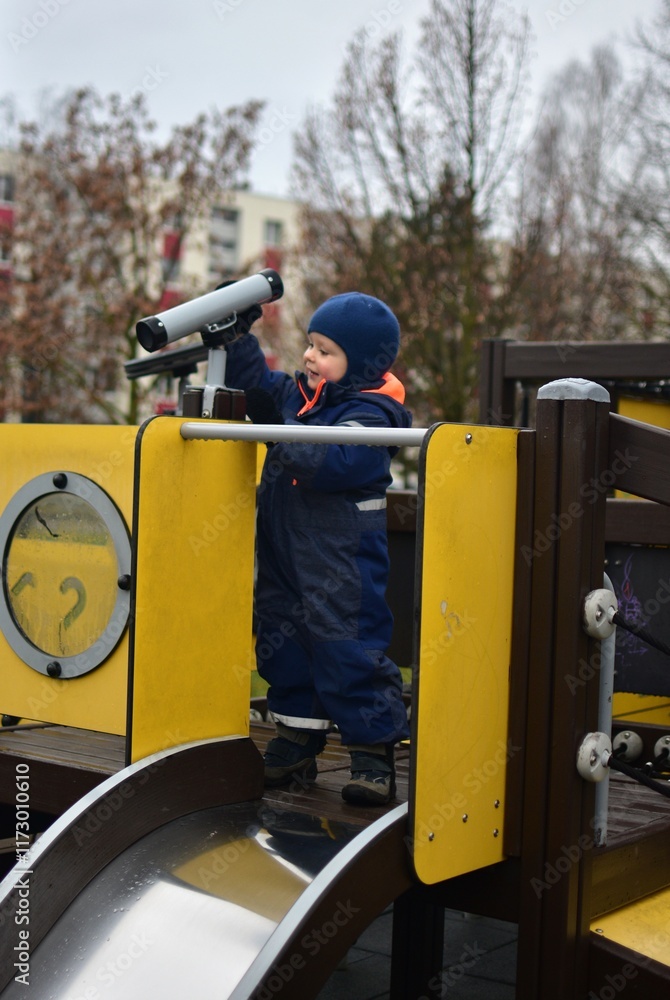 Toddler boy is playing with toy telescope in public playground in cold weather winter time