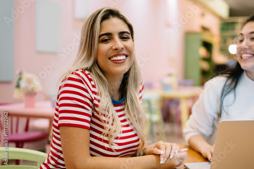 Portrait of cheerful Hispanic hipster girl laughing at camera during collaborative meeting with blurred freelance colleague, happy female students 20 years old enjoying e learning togetherness photo