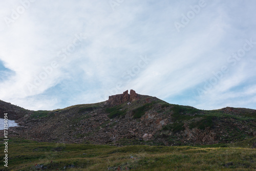 Dramatic view to gate shaped rock outliers on grassy stony hill with lush alpine flora under spindrift clouds in blue sky. Unusual old sharp rocky outlier of star gate shape in center of hill top. photo