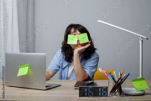 A tired woman with curly hair in a striped shirt sits at a cluttered desk with sticky notes covering her eyes. She rests her head on her hand, surrounded by a laptop, clock, lamp, and stationery.
