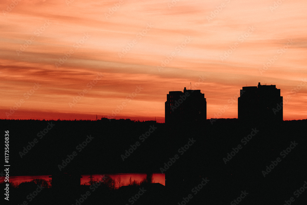Silhouette of two tall buildings against a dramatic orange-red sunset sky with streaks of clouds. The lower part of the image is dark, with reflections of the sunset visible on a body of water. Silhouette of two tall buildings against a dramatic orange-red sunset sky with streaks of clouds. The lower part of the image is dark, with reflections of the sunset visible on a body of water.