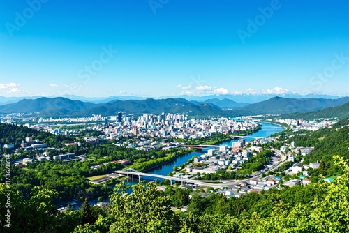 Aerial view of a cityscape surrounded by greenery, mountains, and a river under a bright blue sky. photo