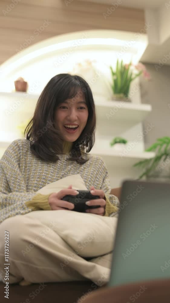 Vertical video of Smiling asian woman enjoying cozy gaming at home, holding controller surrounded by plants in stylish indoor space. Vertical video of Smiling asian woman enjoying cozy gaming at home, holding controller surrounded by plants in stylish indoor space.