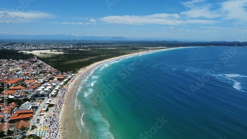 Aerial drone view of Peró Beach, Cabo Frio, Rio de Janeiro, Brazil, during summer, bustling with colorful umbrellas, people, and vibrant beach life under the sunny sky.