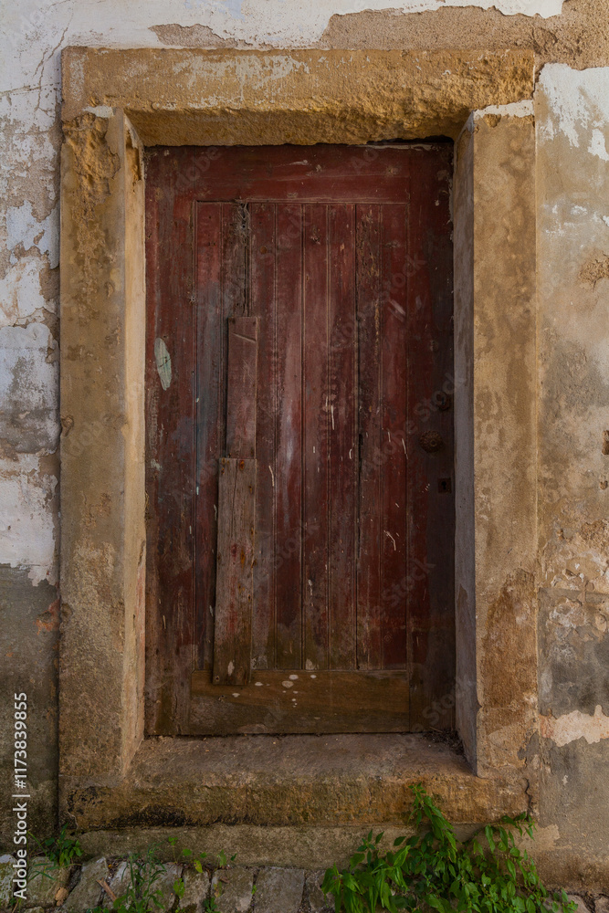 Fototapeta premium A weathered, dark red wooden door with a stone frame is set into a crumbling wall, with weeds growing at the base