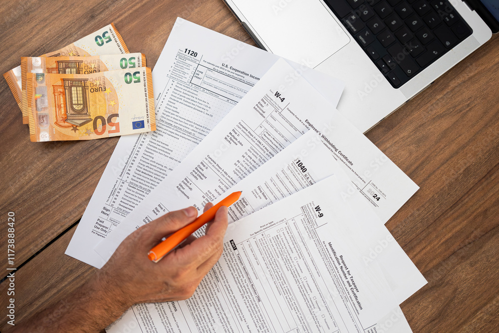 Fototapeta premium Close-up of a man filling out tax forms with euro banknotes and a laptop on a wooden table. Tax preparation and financial management concept. Ideal for tax season and accounting visuals