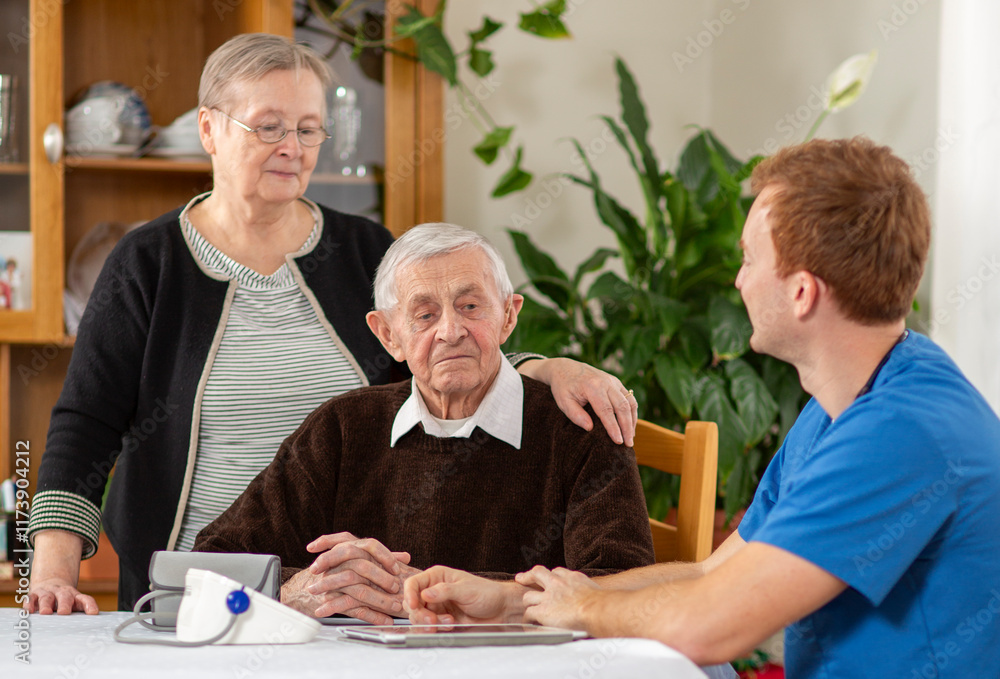 Elderly man seated at a table, accompanied by a healthcare professional in blue scrubs and a woman standing beside him, creating a caring and supportive atmosphere