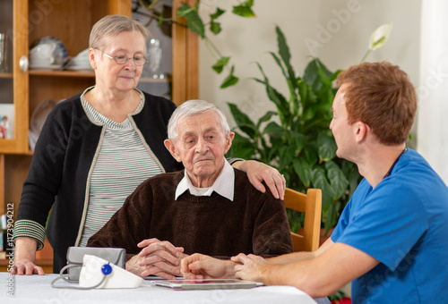 Obraz na płótnie Elderly man seated at a table, accompanied by a healthcare professional in blue scrubs and a woman standing beside him, creating a caring and supportive atmosphere w salonie