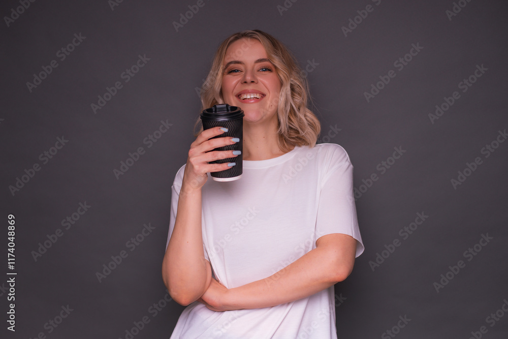 Smiling woman holding a black takeaway coffee cup, wearing a white t-shirt, standing against a gray background, exuding confidence and positivity in a minimalist lifestyle scene. Smiling woman holding a black takeaway coffee cup, wearing a white t-shirt, standing against a gray background, exuding confidence and positivity in a minimalist lifestyle scene.
