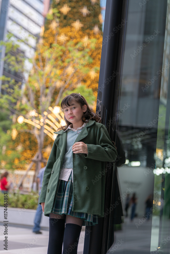 Obraz premium A young girl in a green coat posing gracefully in a festive urban setting, with a glowing Christmas tree and sparkling lights in the background, capturing a serene and joyful holiday moment.
