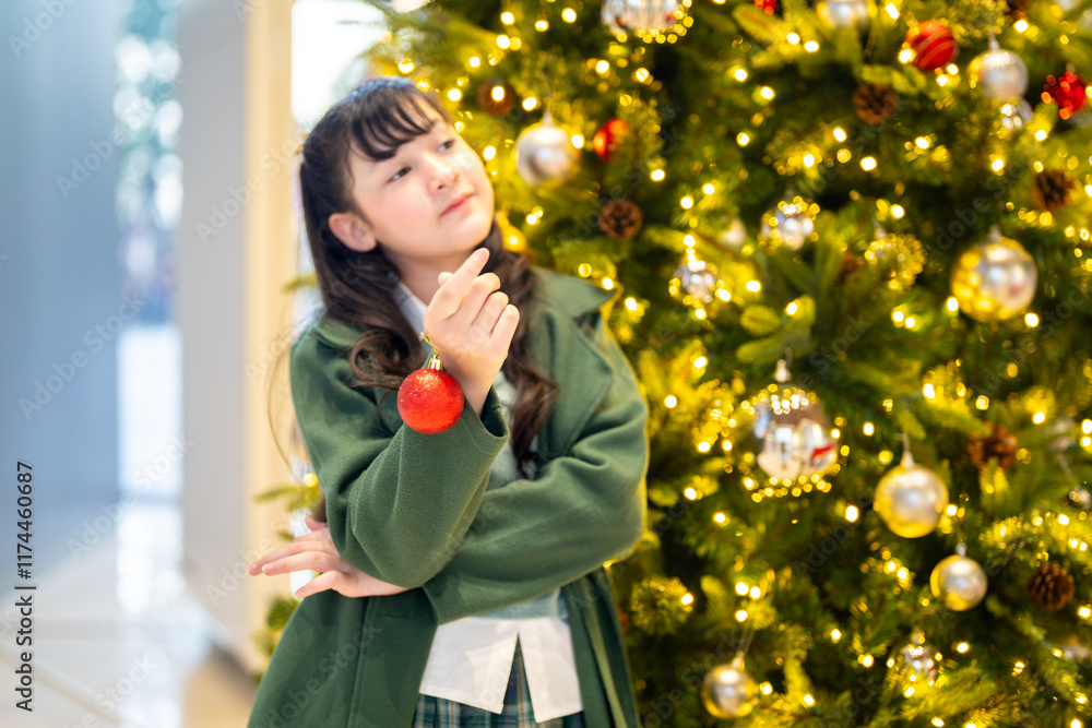 A young girl in a green coat playfully holding a red Christmas ornament near her ear, standing beside a beautifully decorated Christmas tree adorned with lights and festive ornaments. A young girl in a green coat playfully holding a red Christmas ornament near her ear, standing beside a beautifully decorated Christmas tree adorned with lights and festive ornaments.