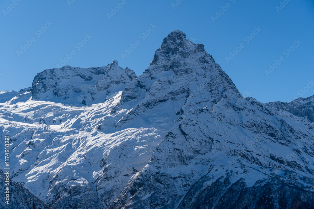 Fototapeta premium Dombay. Karachay-Cherkessia. Giant rocks with snow-capped peaks and glaciers rise majestically against blue autumn sky. Pine and spruce forests grow at foot of mountains. Sunny day. Autumn 2024