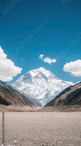 A majestic, snow-capped mountain peak under a clear blue sky, surrounded by rocky terrain and scattered clouds.