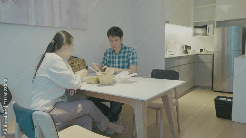 A relaxed dining moment in a contemporary kitchen setting, featuring two people enjoying their meal at a light-colored wooden table. The ambiance is cozy and inviting, showcasing a minimalist design.