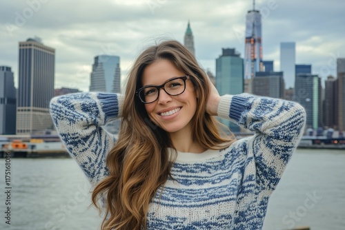 cute, smiling brunette girl with glasses posing in front of buildings and river at Manhattan background wearing blue white pattern sweater. The city skyline is visible behind her
