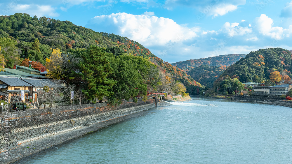 Fototapeta premium A picturesque view of the Uji Riverbank with lush green trees and autumn foliage. Traditional Japanese buildings line the river. Kyoto, Japan.
