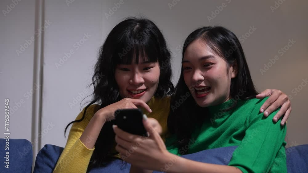 Young female friends enjoying lighthearted smartphone moment, sitting together on home sofa and sharing digital content with genuine laughter and connection Young female friends enjoying lighthearted smartphone moment, sitting together on home sofa and sharing digital content with genuine laughter and connection