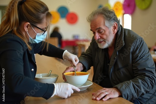 A woman in gloves and a mask is serving soup to a disheveled, dirty homeless man at a community center. The background shows a warm room with colorful decorations and tables photo