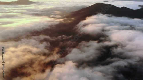 Aerial view of a mountain peak surrounded by morning fog and glowing clouds during sunrise, creating a mystical and serene landscape.