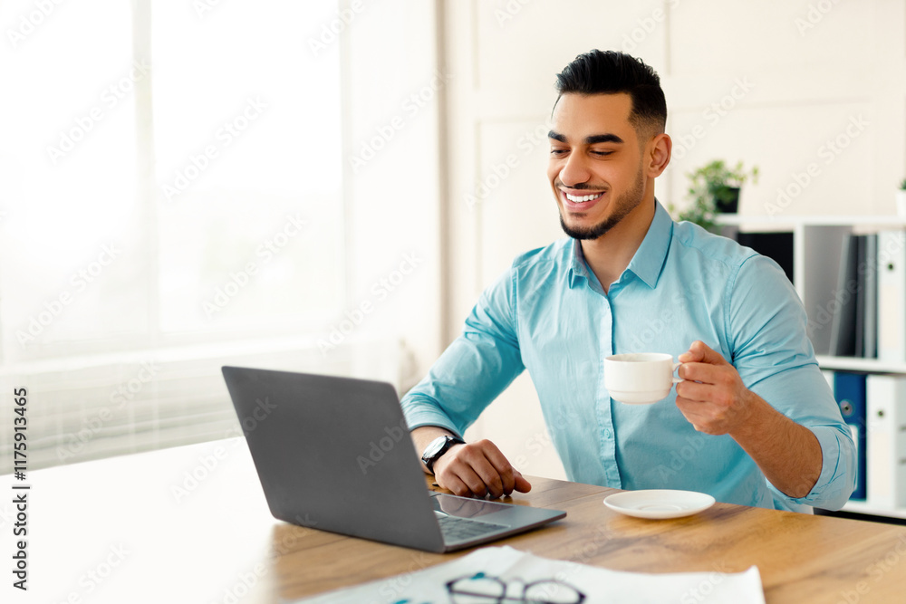 Happy young Arab man drinking coffee while using laptop at modern office. Handsome Eastern entrepreneur enjoying hot drink during online meeting with partner or colleague Happy young Arab man drinking coffee while using laptop at modern office. Handsome Eastern entrepreneur enjoying hot drink during online meeting with partner or colleague