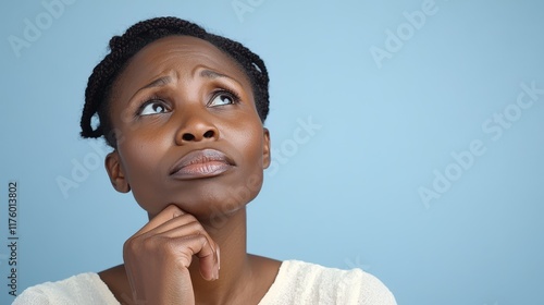 A pensive woman gazes upward, lost in thought, resting her chin on her hand against a soft blue background. photo