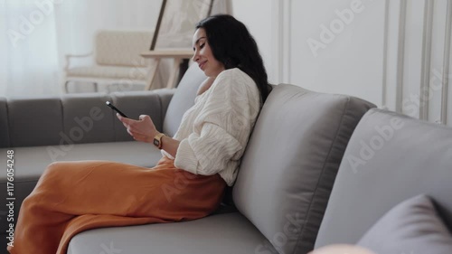 A woman is comfortably seated on the couch in the house, using her cell phone