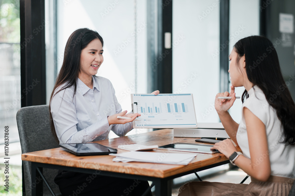Two businesswomen discussing financial charts on a tablet in a bright, modern office environment. Two businesswomen discussing financial charts on a tablet in a bright, modern office environment.