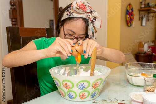 Young woman with Down syndrome, wearing glasses, breaks an egg into a bowl full of flour. She wears an apron, concentrating on her culinary activity, enjoying the cooking process. Trisomy 21