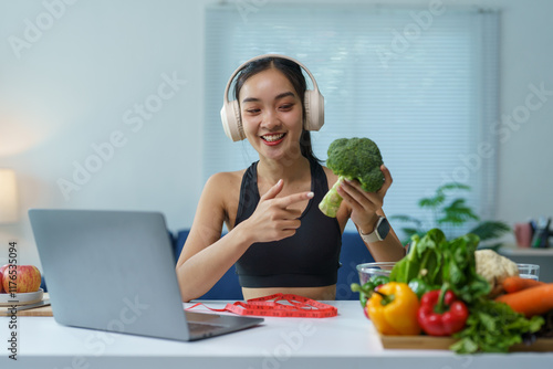 Asian nutritionist wearing headphones and holding broccoli while giving online healthy eating consultation via video call with laptop at home photo