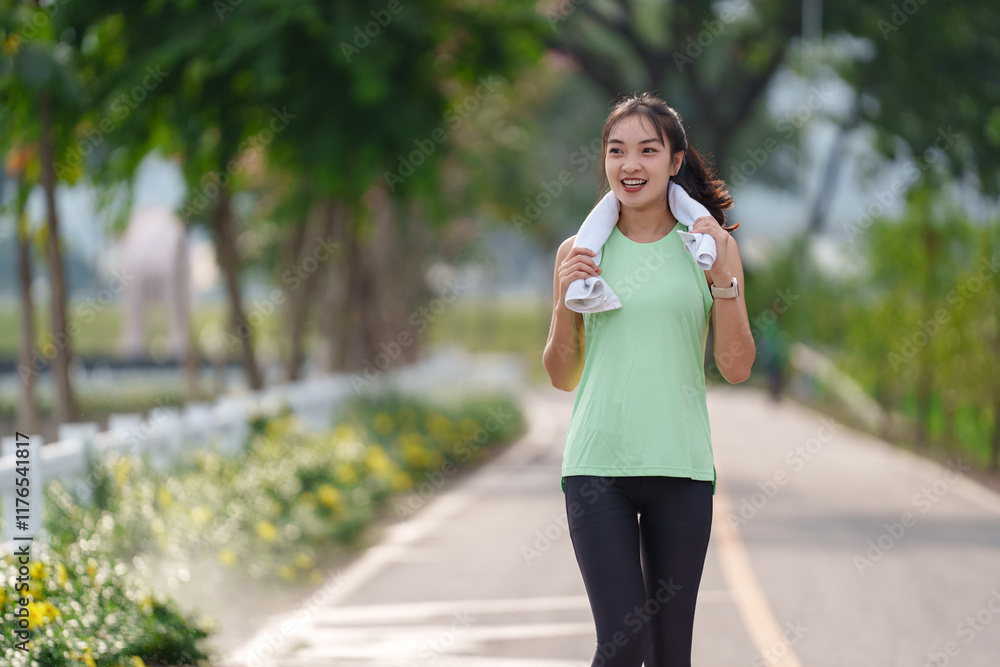 Smiling young asian woman walking through a park after exercising, holding a white towel around her neck, enjoying the beauty of nature on a sunny day Smiling young asian woman walking through a park after exercising, holding a white towel around her neck, enjoying the beauty of nature on a sunny day