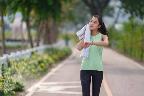 Young sporty asian woman wiping sweat with a towel while checking the time on her smartwatch after an invigorating run in the park, enjoying the fresh air and sunshine