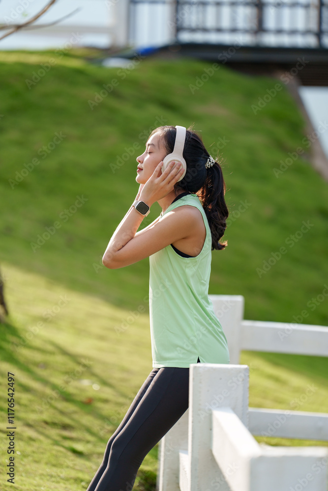 Young asian sportswoman wearing wireless headphones and smartwatch, enjoying music while relaxing in a park, leaning on a white wooden fence, after exercising